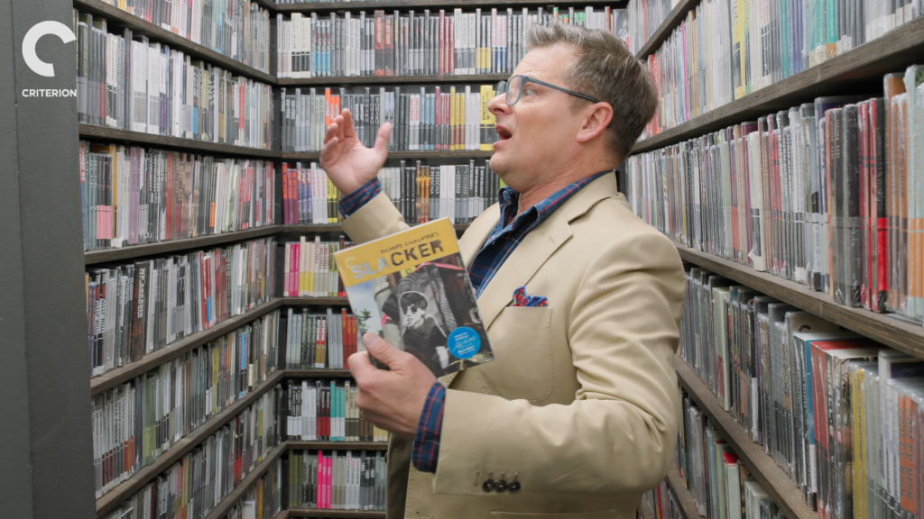 Man in a beige blazer and plaid shirt holding a magazine titled 'SLACKER' in a narrow aisle lined with shelves of books or magazines; his hand is raised in a gesturing pose, and a Criterion logo is visible on the left.