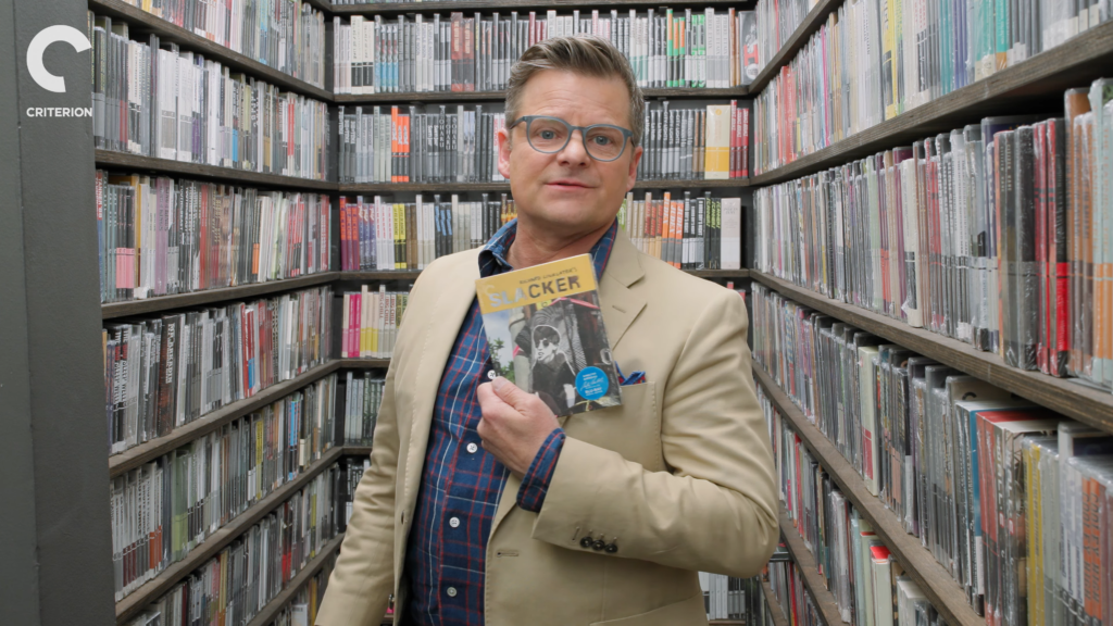 Man in a beige blazer and glasses holds a magazine titled 'Slacker' in a narrow library aisle.