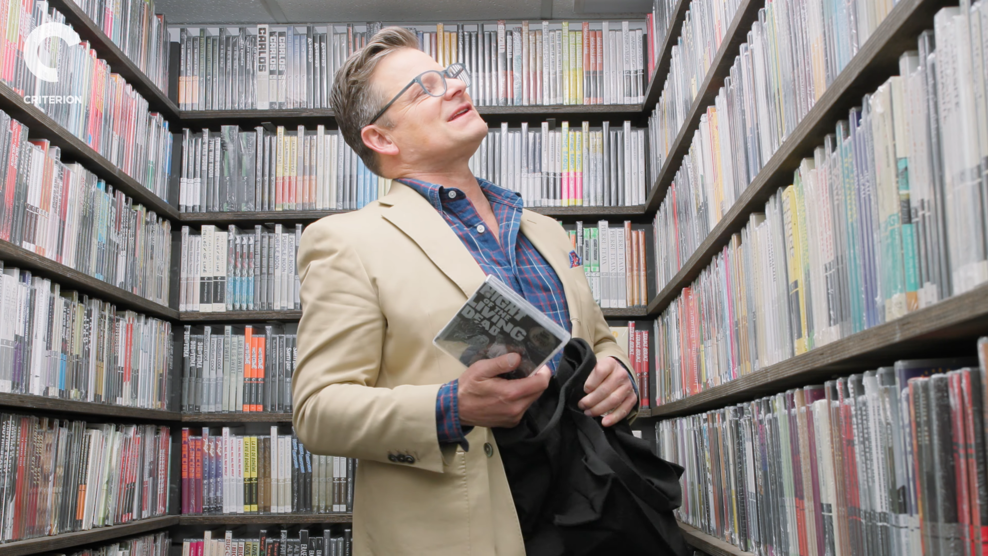 Man in a beige blazer and plaid shirt stands in a library aisle, looking up, holding a DVD case.
