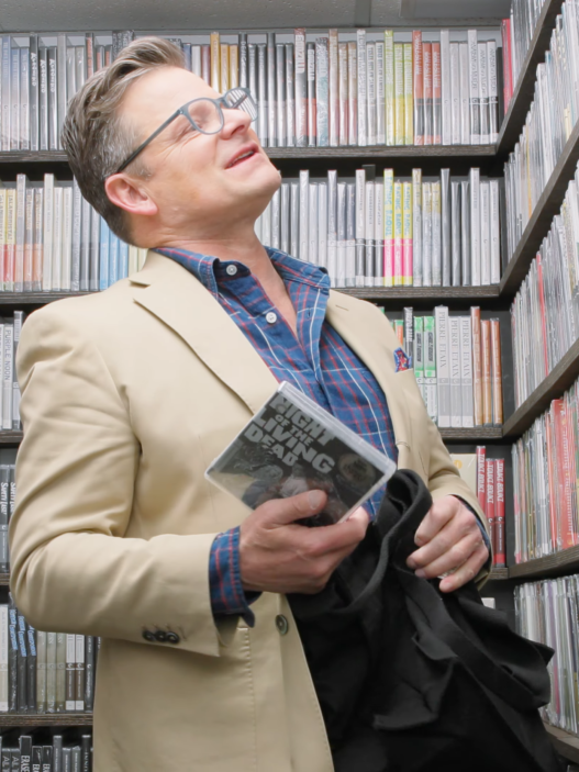 Man in a beige blazer and plaid shirt stands in a library aisle, looking up, holding a DVD case.