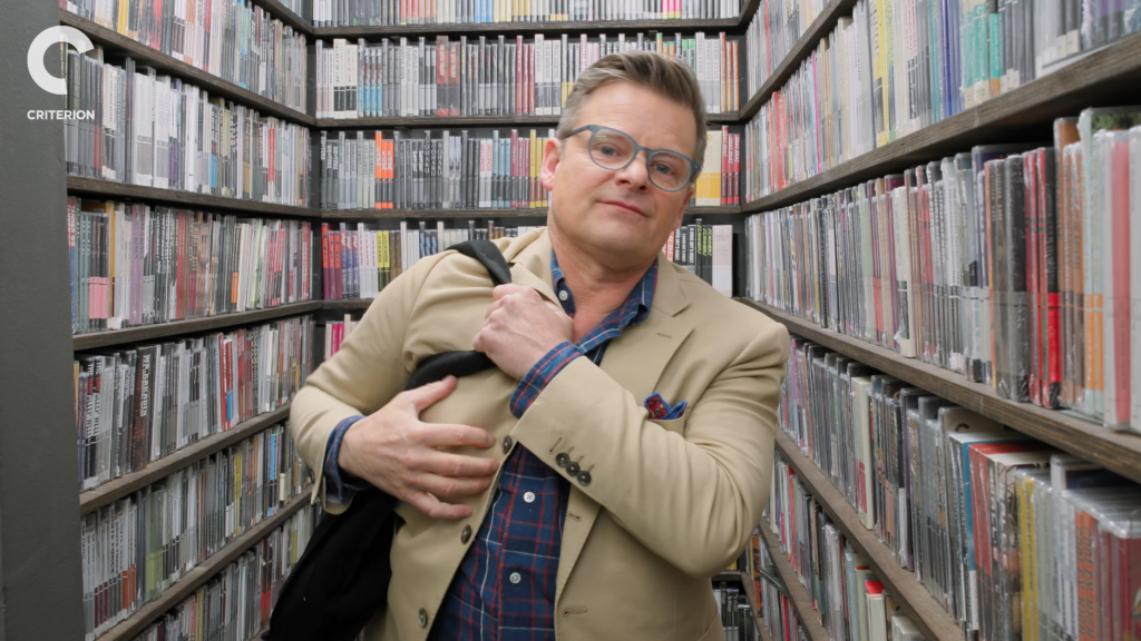 Man in a tan blazer and glasses, holding a black coat, posing in a narrow aisle surrounded by colorful magazine shelves in a library/record store setting.