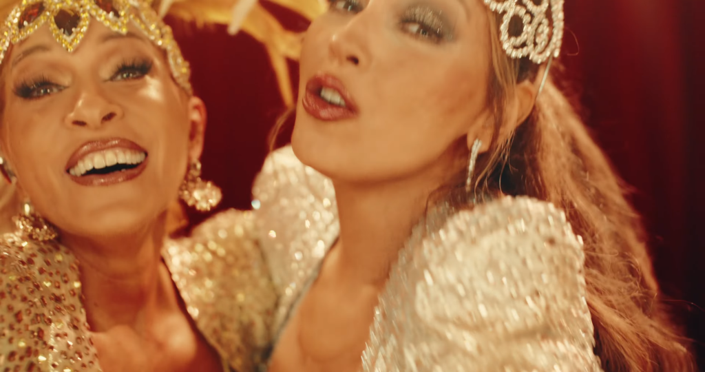 Two women wearing gold jeweled crowns and glittery outfits, smiling at a festive event.