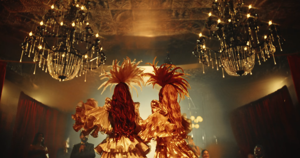 Dancers in gold costumes with tall feather headdresses perform on a dimly lit stage under crystal chandeliers.