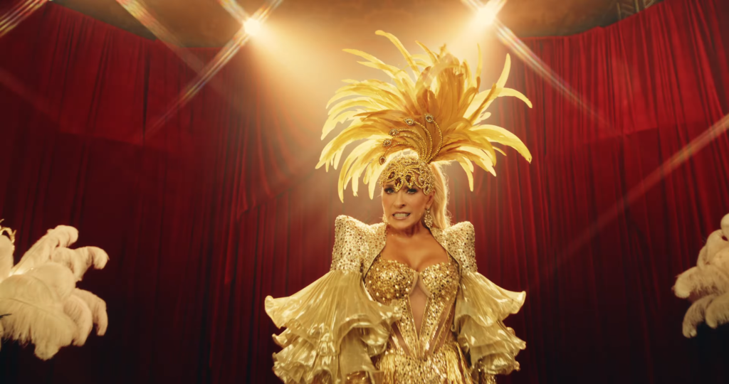 Showgirl in a gold sequined costume and towering feather headdress on a stage with red curtains under bright lights.