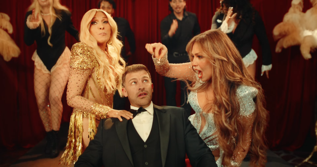Man in a black tuxedo sits as two glamorous women in gold and silver sequined dresses lean in beside him on a stage with a red curtain backdrop.