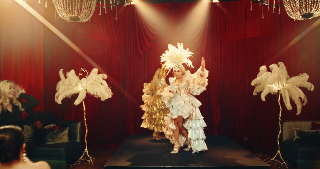 Dancers in sequined showgirl costumes with white feather headdresses performing on a red-curtained stage under warm spotlights