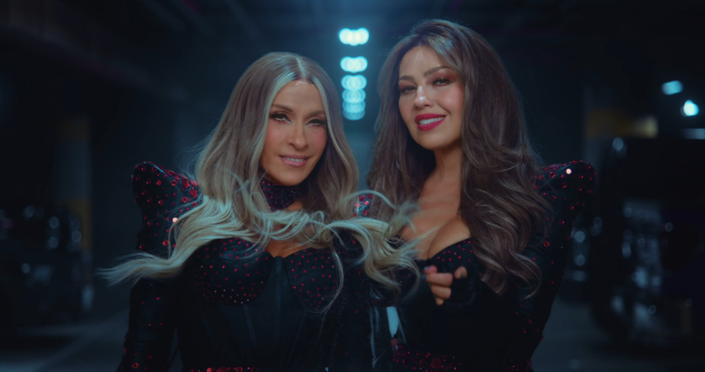 Two women in matching black outfits with red sequins pose and smile in a dim backstage setting.