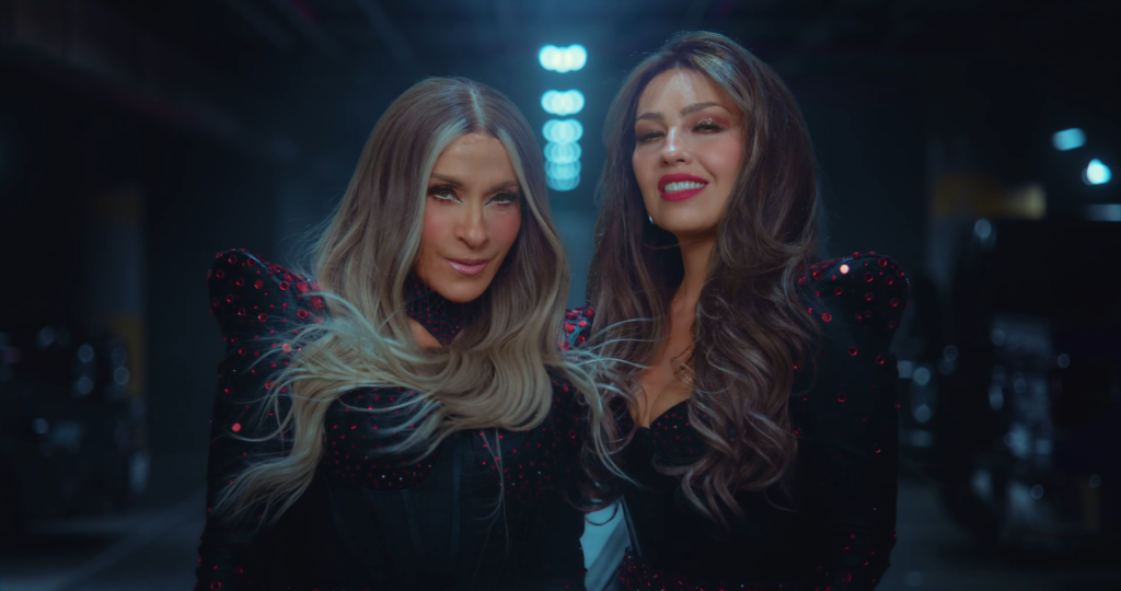 Two women in sparkly black outfits with red sequins posing and smiling in a dim backstage area with blue lighting behind them.