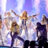 Pop star center stage in a metallic blue bodysuit, arms outstretched, flanked by dancers in silver outfits on a glittering stage with fireworks/pyrotechnics behind.