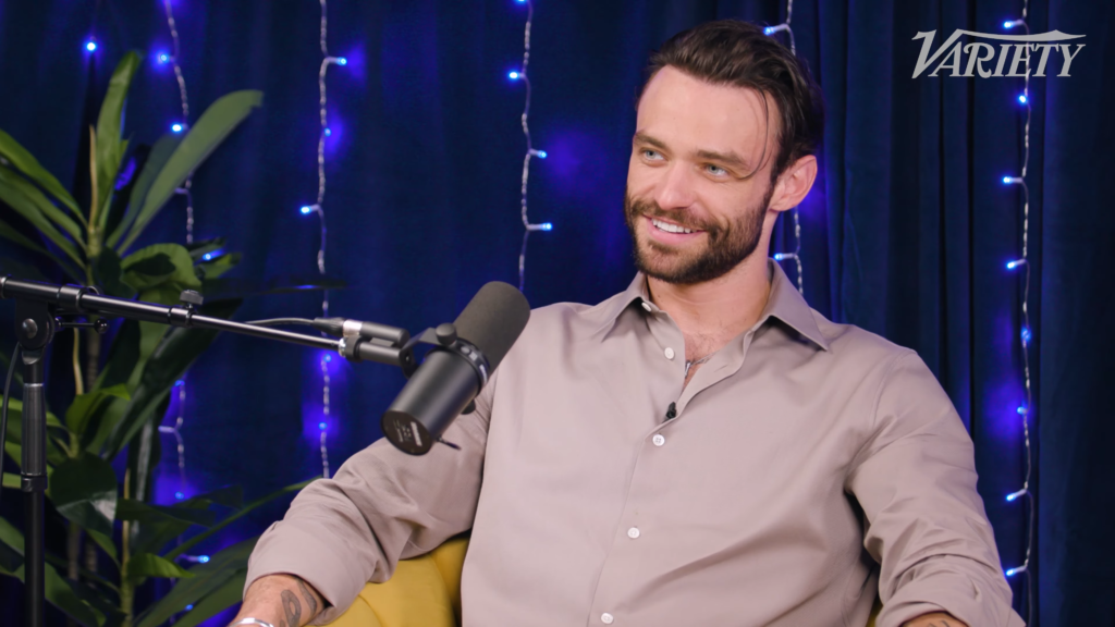Man with beard in a light gray shirt sits at a podcast microphone, smiling, with blue string lights and a Variety logo visible in the corner.