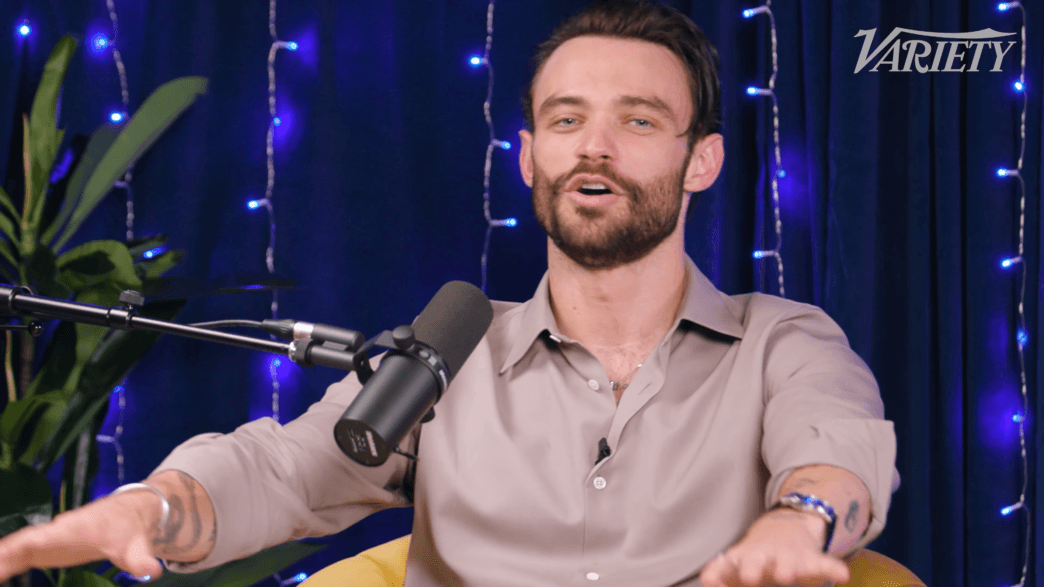 Male podcast host speaking into a microphone, gesturing with hands, blue string lights and Variety logo in the background.