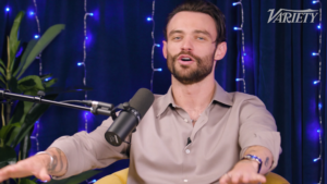 Male podcast host speaking into a microphone, gesturing with hands, blue string lights and Variety logo in the background.