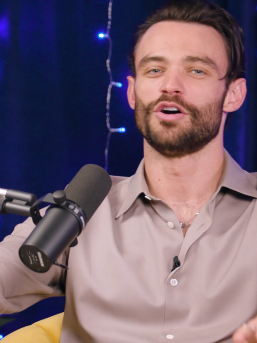 Male podcast host speaking into a microphone, gesturing with hands, blue string lights and Variety logo in the background.