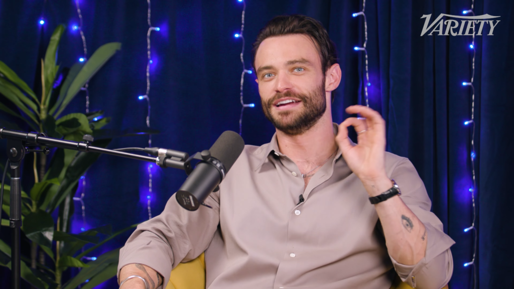Man with a beard speaks into a microphone during a podcast interview, blue-lit studio with string lights and a Variety logo in the corner.