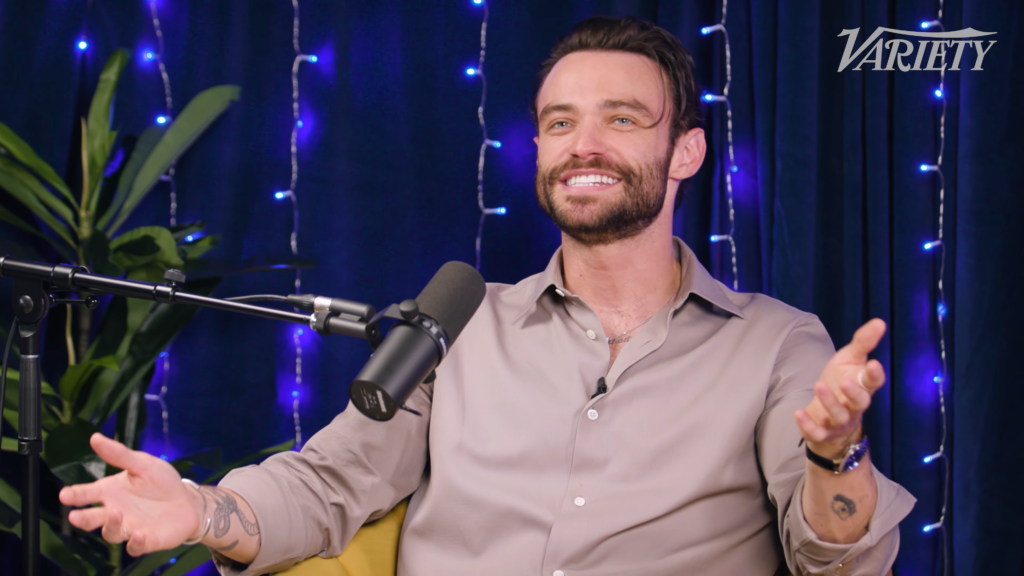 Bearded man in a light shirt speaks into a studio microphone during a podcast, with blue string lights and a Variety logo in the background.
