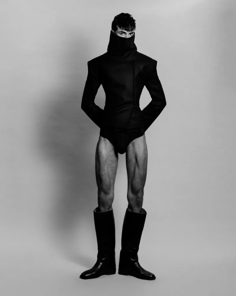 Monochrome fashion shot of a model in a sculptural black coat with a high neck covering the lower face, wearing tall black boots and no visible shirt, standing against a gray backdrop.
