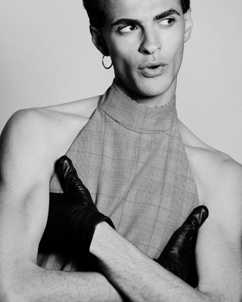 Black-and-white fashion portrait of a model wearing a plaid high-neck halter top with black gloves crossed over the chest, looking to the side.