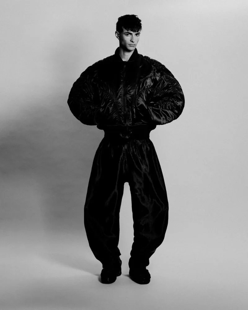 Male model in an oversized black puffer jacket and loose black pants, standing in a studio with a neutral backdrop.