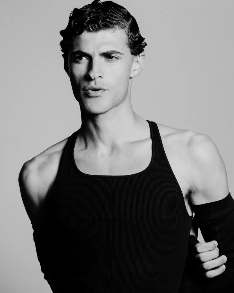 Male model in a black sleeveless shirt, looking to the left, with short wavy hair in a studio portrait.