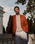 Man leaning on a white balustrade in a rust-orange blazer, beige sweater, and white pants outdoors.