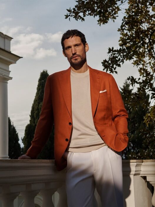 Man leaning on a white balustrade in a rust-orange blazer, beige sweater, and white pants outdoors.