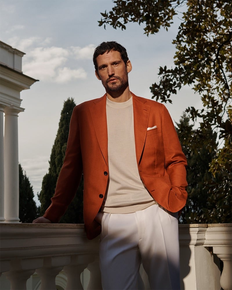 Man leaning on a white balustrade in a rust-orange blazer, beige sweater, and white pants outdoors.