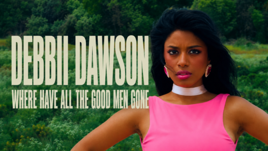 Debbii Dawson stands in a vivid pink dress against a lush green meadow in a cinematic still from “Where Have All The Good Men Gone.”