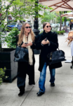 Two stylish women walking on a city sidewalk with black handbags, wearing sunglasses and coats.