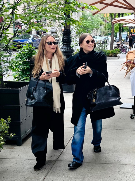 Two stylish women walking on a city sidewalk with black handbags, wearing sunglasses and coats.