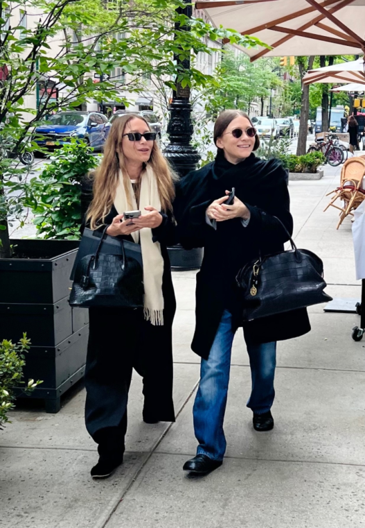 Two stylish women walking on a city sidewalk with black handbags, wearing sunglasses and coats.