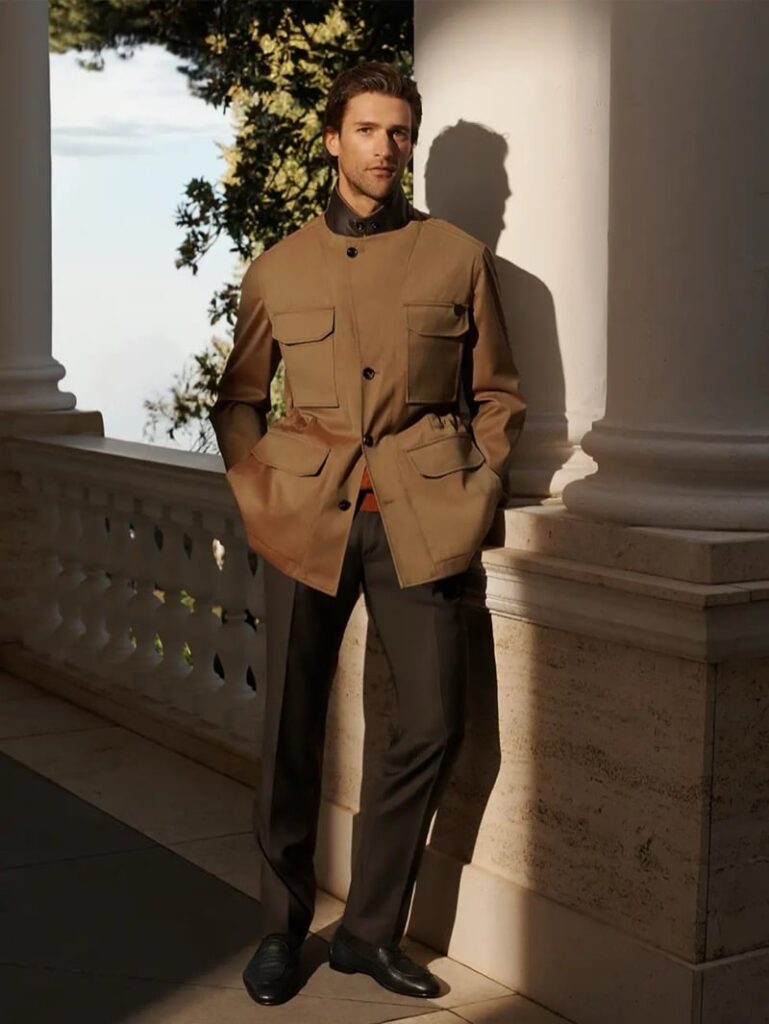 Man in a tan utility jacket with large front pockets stands with hands in pockets beside marble columns on a sunny terrace.