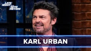 Smiling male guest on a late-night talk show, with 'KARL URBAN' text overlay and blue neon horizontal lines across the frame.