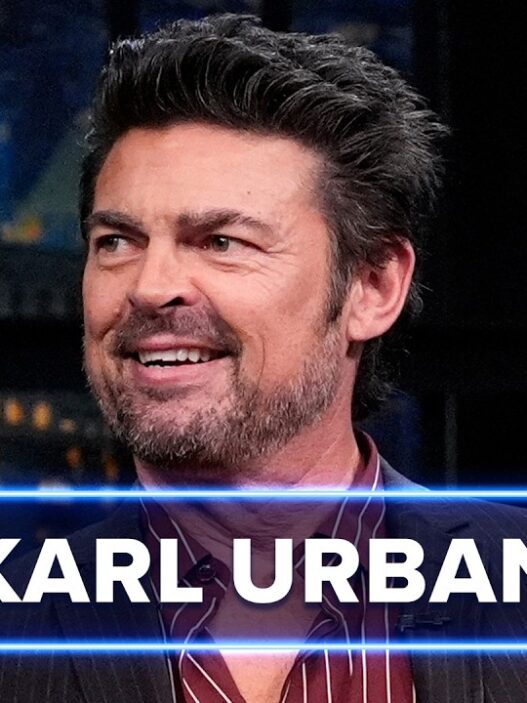 Smiling male guest on a late-night talk show, with 'KARL URBAN' text overlay and blue neon horizontal lines across the frame.