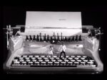Giant vintage typewriter on a stage in black and white, with performers standing on the keys and a large sheet of paper behind them.