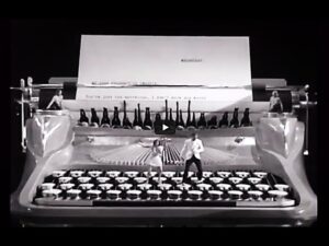 Giant vintage typewriter on a stage in black and white, with performers standing on the keys and a large sheet of paper behind them.