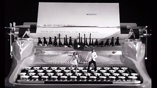 Giant vintage typewriter on a stage in black and white, with performers standing on the keys and a large sheet of paper behind them.