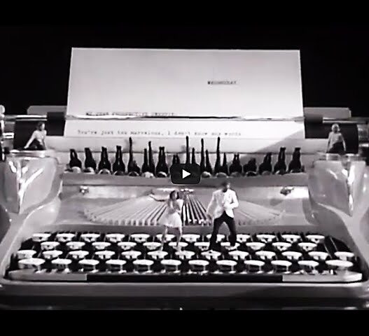 Giant vintage typewriter on a stage in black and white, with performers standing on the keys and a large sheet of paper behind them.