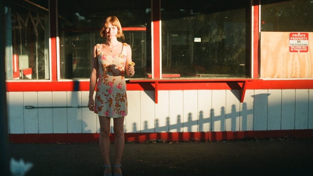 Frankie June standing in warm sunlight outside a red-and-white storefront holding an ice cream cone