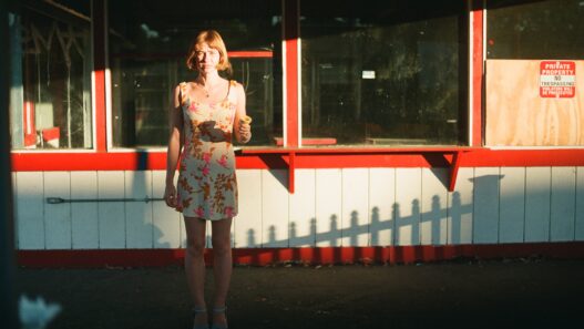 Frankie June standing in warm sunlight outside a red-and-white storefront holding an ice cream cone