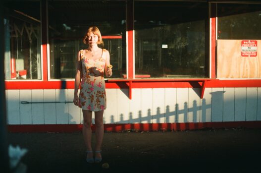 Frankie June standing in warm sunlight outside a red-and-white storefront holding an ice cream cone