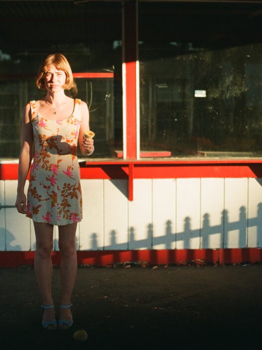 Frankie June standing in warm sunlight outside a red-and-white storefront holding an ice cream cone