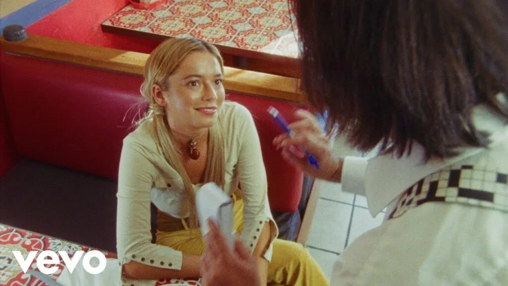 Tele seated inside a retro diner while speaking with another person in the Honest Man music video