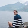 Federico Cola sits beside Lake Como in a navy striped sweater with mountains behind him