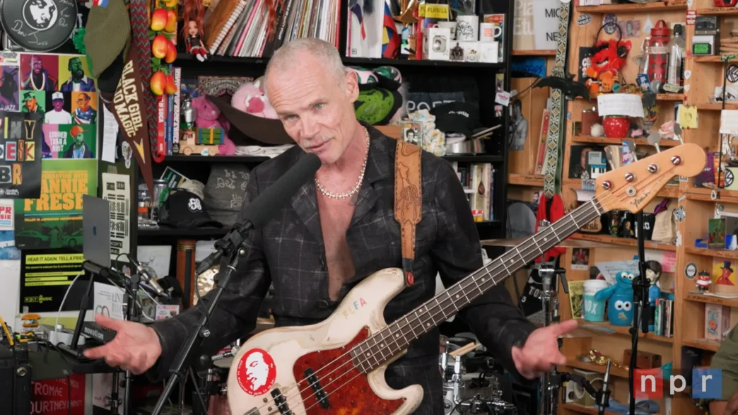 Flea speaking at the NPR Tiny Desk with his bass strapped across his chest during the Honora Band performance