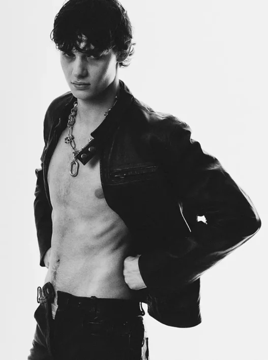 Juan Navarro poses in a black-and-white fashion portrait wearing an open leather jacket, chain necklace and dark trousers.