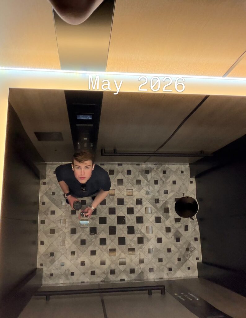 Iain Armitage looking up toward an elevator mirror while holding a camera in May 2026