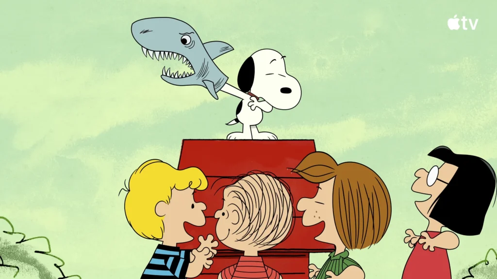 Snoopy standing on his red doghouse with a shark puppet as the Peanuts gang watches
