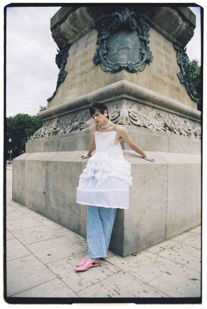 Ruslan leaning against a stone monument in a white hoop-skirt top, blue trousers and pink shoes