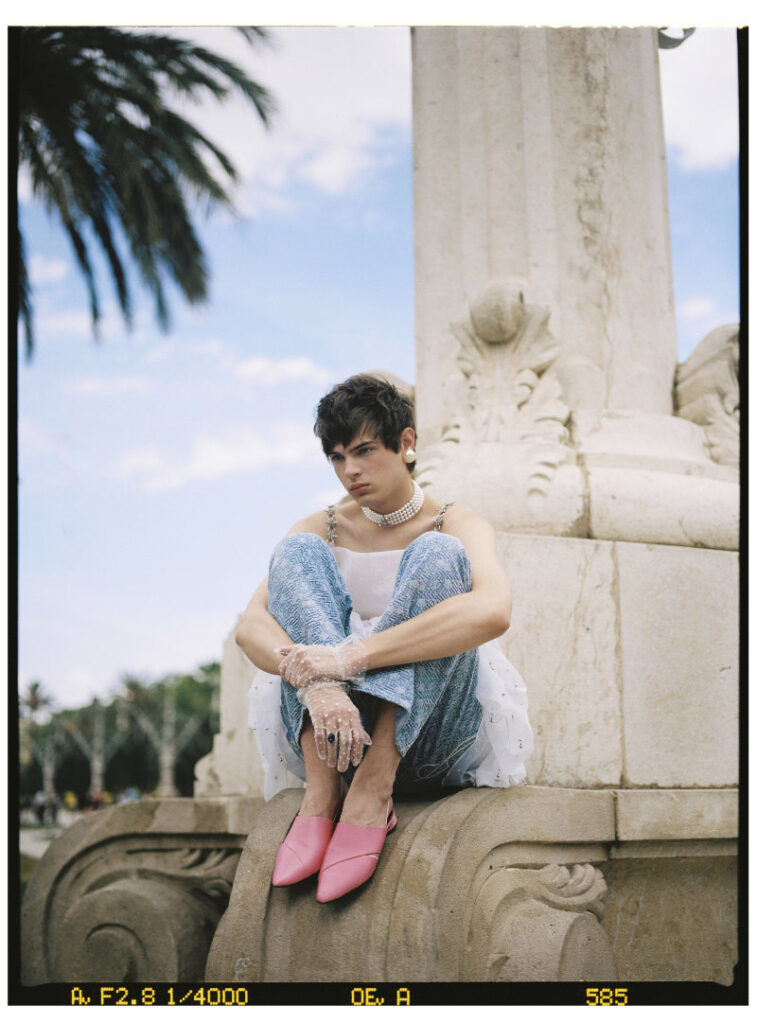 Ruslan seated on a stone monument in a white sculptural top, blue patterned trousers and pink shoes
