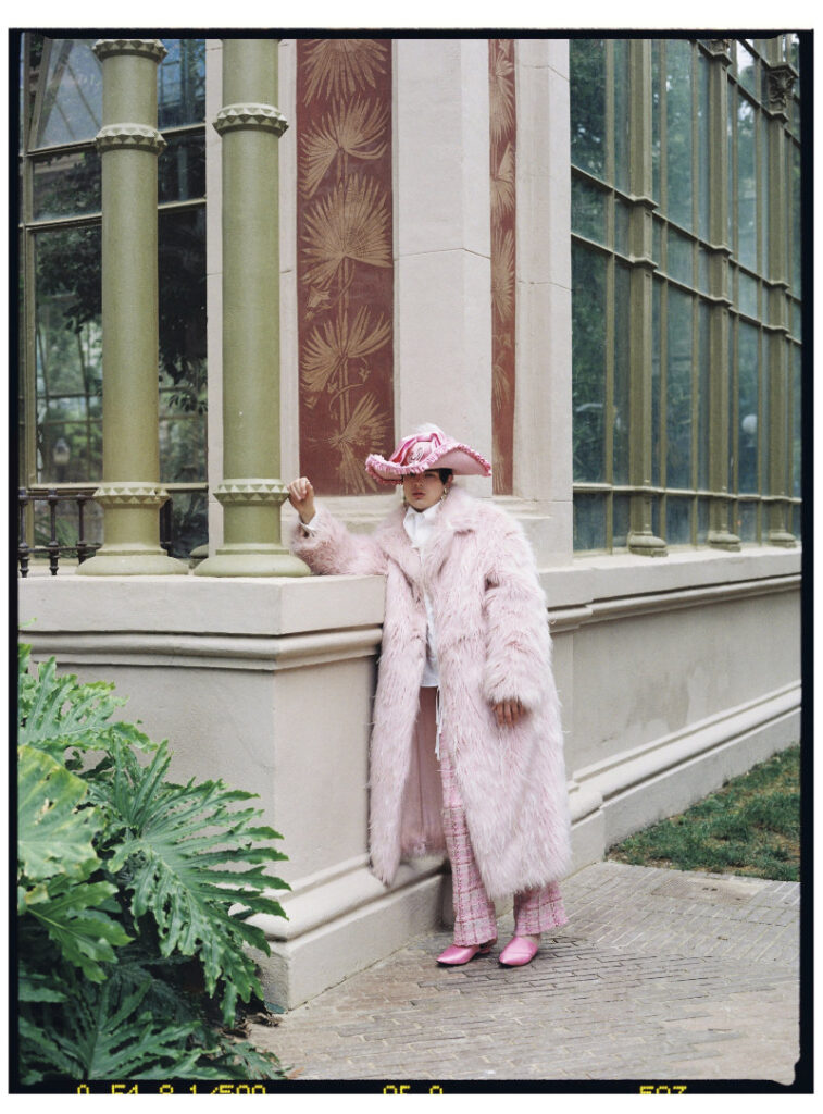 Ruslan wearing a long pink faux-fur coat, ruffled hat, pink trousers and shoes outside a glass pavilion